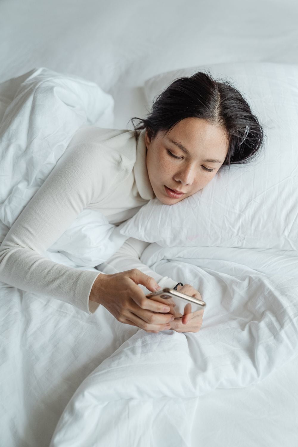 A woman lying in bed with white sheets and pillows looks at her smartphone, appearing relaxed and comfortable.