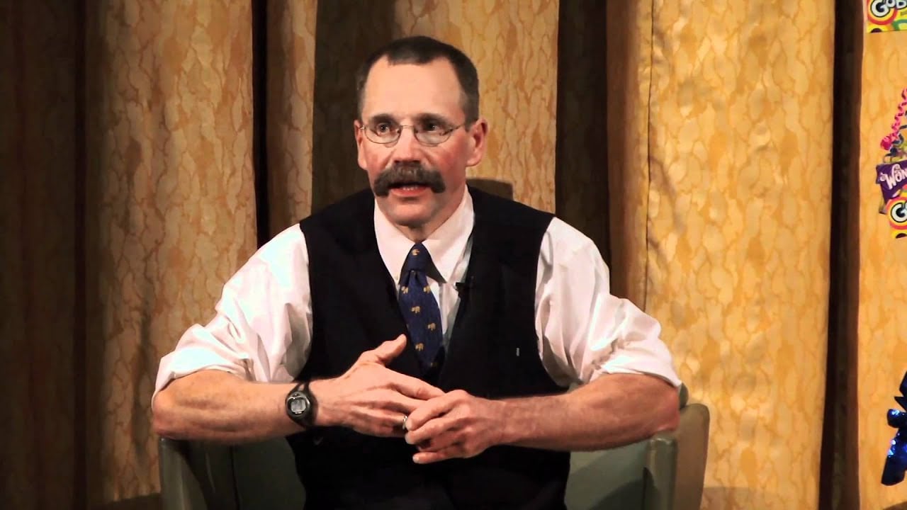 A man with a mustache, glasses, and short hair sits in an armchair. He is wearing a white shirt, dark vest, tie, and watch, gesturing with his hands. The background features gold patterned curtains.