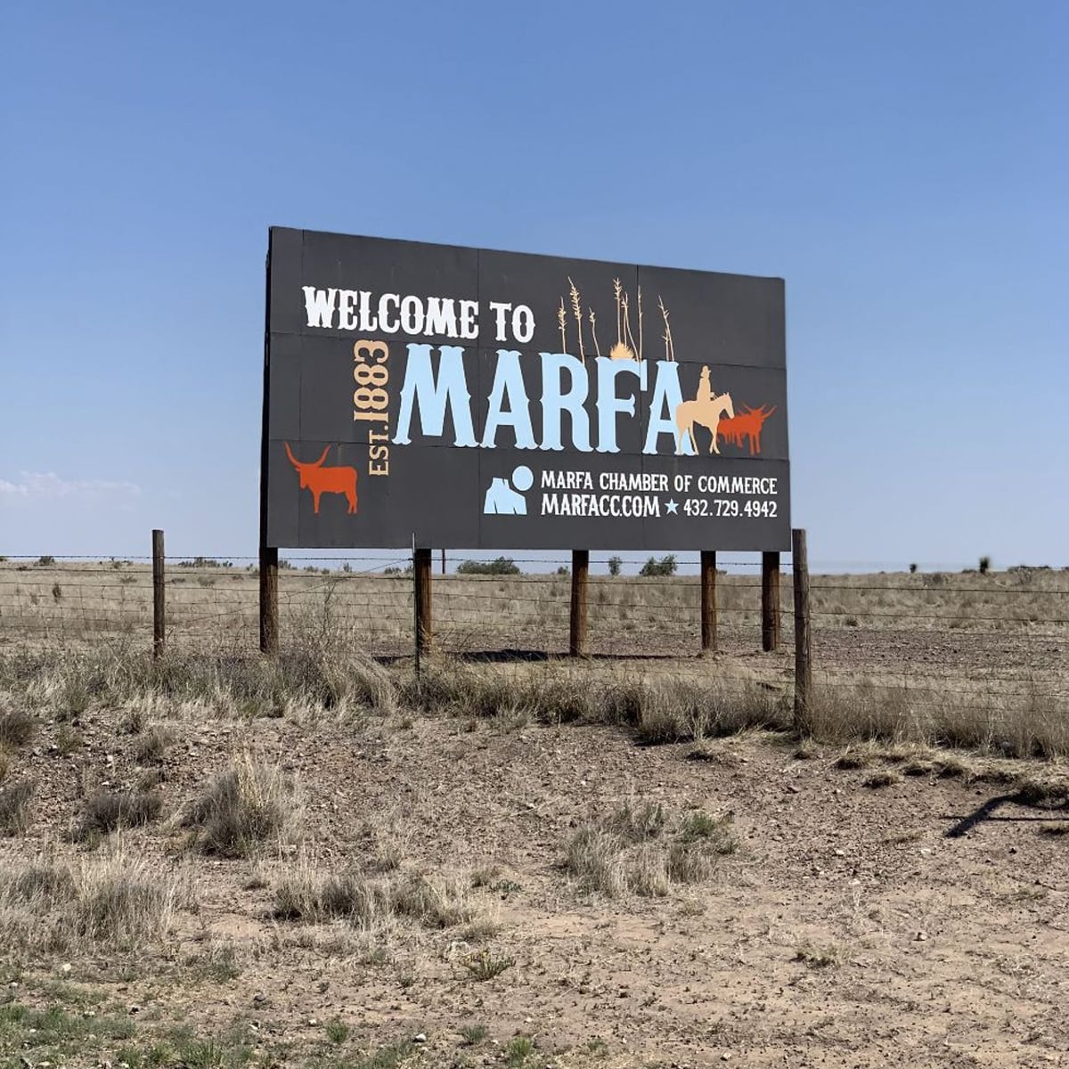 A roadside billboard in a dry, grassy field reads, "Welcome to Marfa Est. 1883," with images of a longhorn, a pronghorn, and the Marfa Chamber of Commerce contact details.