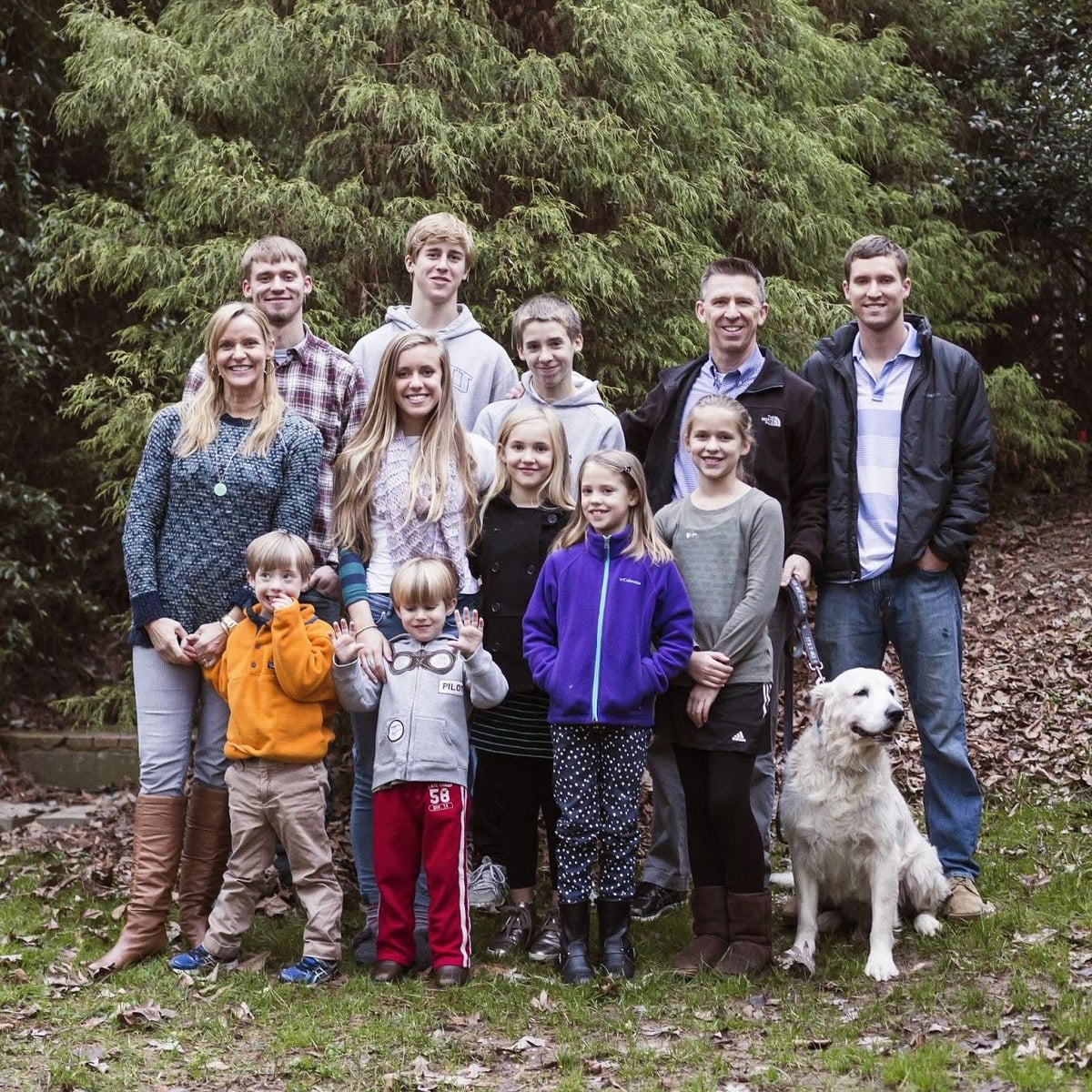 A group of thirteen people, including adults and children, and a white dog pose together outdoors on grass with trees and fallen leaves in the background. Everyone is smiling and dressed in casual clothing.
