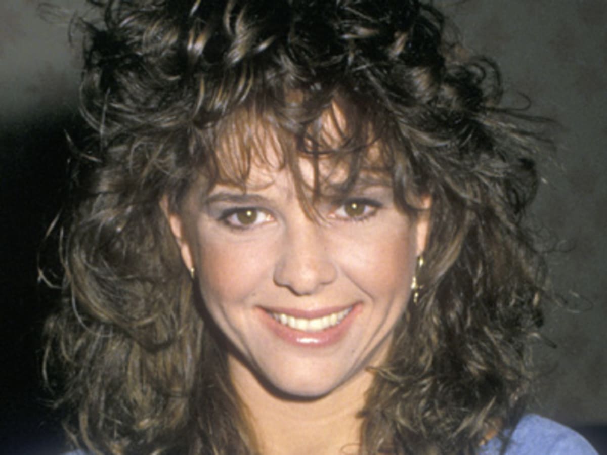 A woman with curly brown hair and bangs smiles at the camera. She is wearing a light blue top and small hoop earrings. The background is neutral and out of focus.