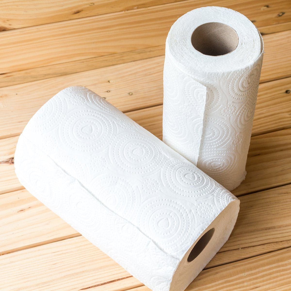 Two rolls of white paper towels with embossed circular patterns are placed on a light wooden surface. One roll is standing upright, while the other is lying down.
