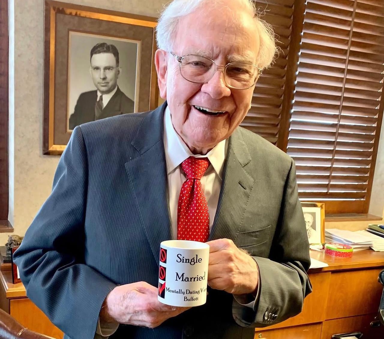 An elderly man in a suit and red tie smiles while holding a mug that reads "Single, Married, Mentally Dating Warren Buffett" in an office with wooden blinds and framed photos on the wall behind him.