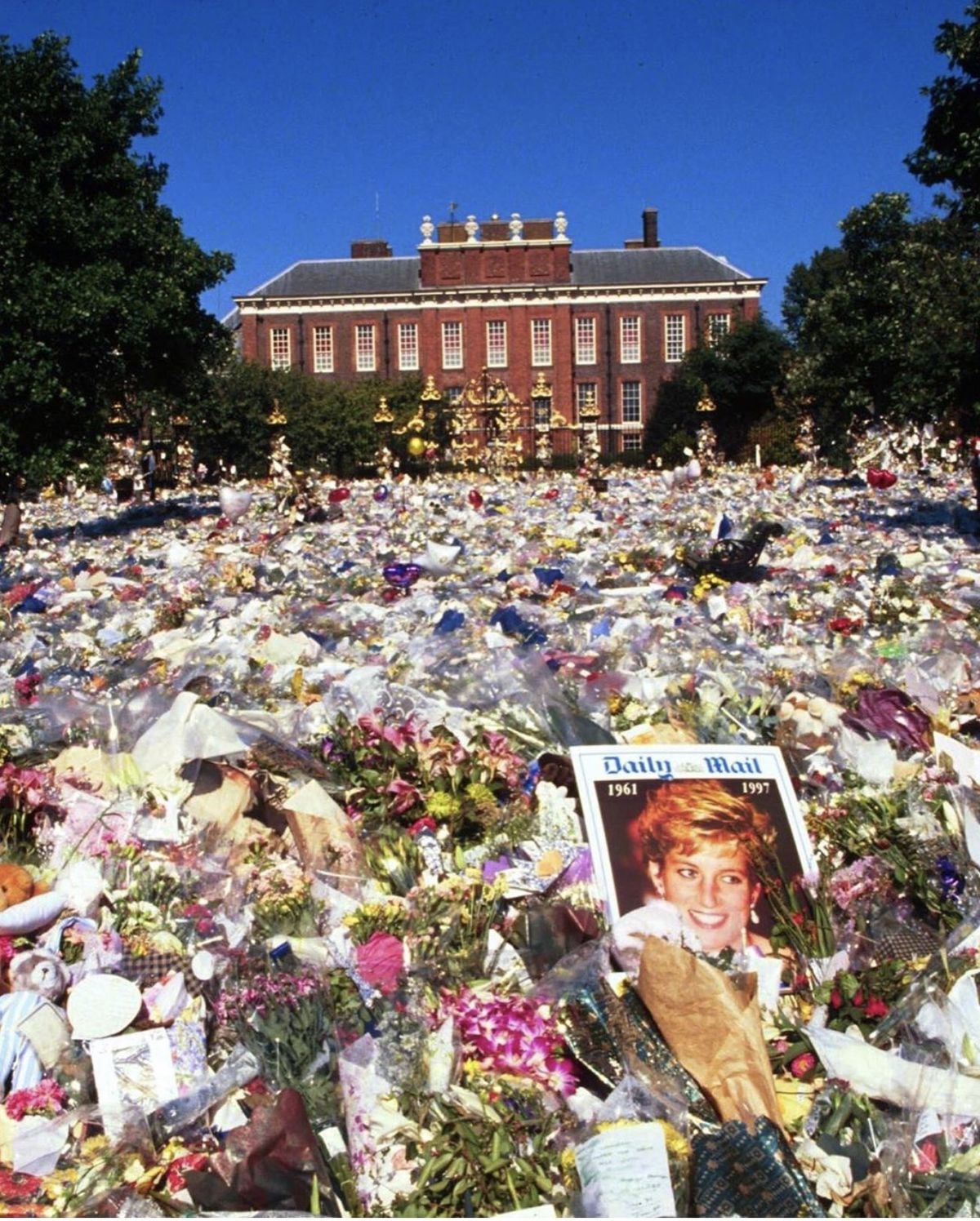 A vast sea of flowers, cards, and tributes covers the ground outside a large brick building, with a prominent photograph and newspaper featuring Princess Diana visible among the memorial items.