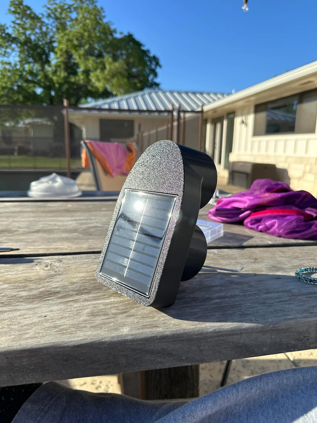 A small black solar-powered device with a reflective front sits on a wooden outdoor table. In the background, there is laundry drying, a fence, and a house with a metal roof under a clear blue sky.