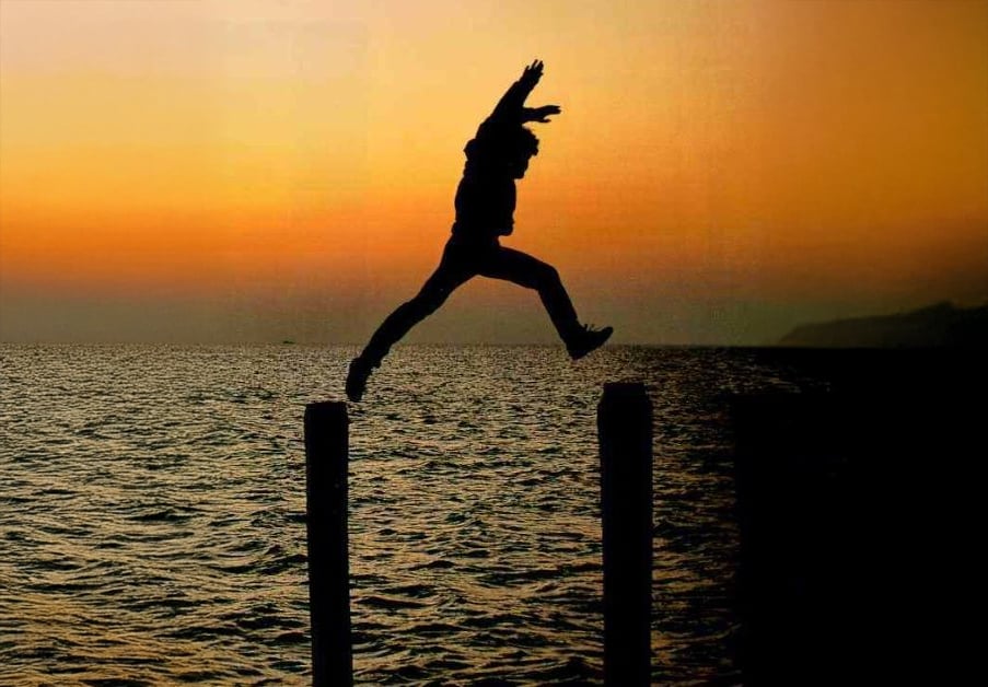 Silhouette of a person leaping between two wooden posts over water at sunset, with an orange sky and distant hills visible in the background.