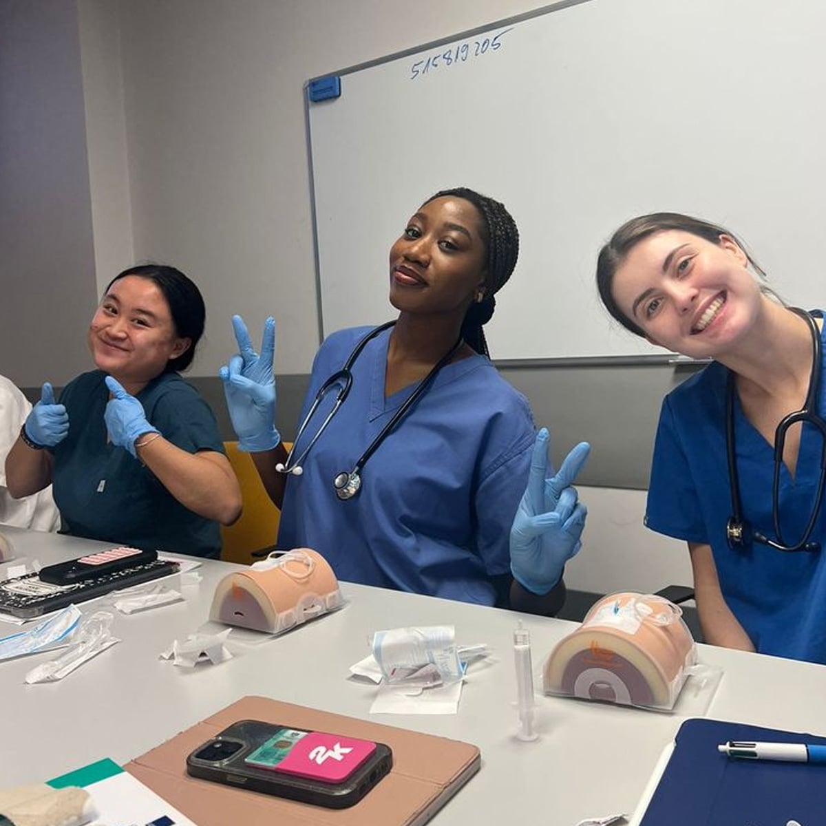 Three medical students in scrubs and gloves sit at a table with medical training models, smiling and making peace signs. Medical supplies and a phone are on the table; a whiteboard is in the background.