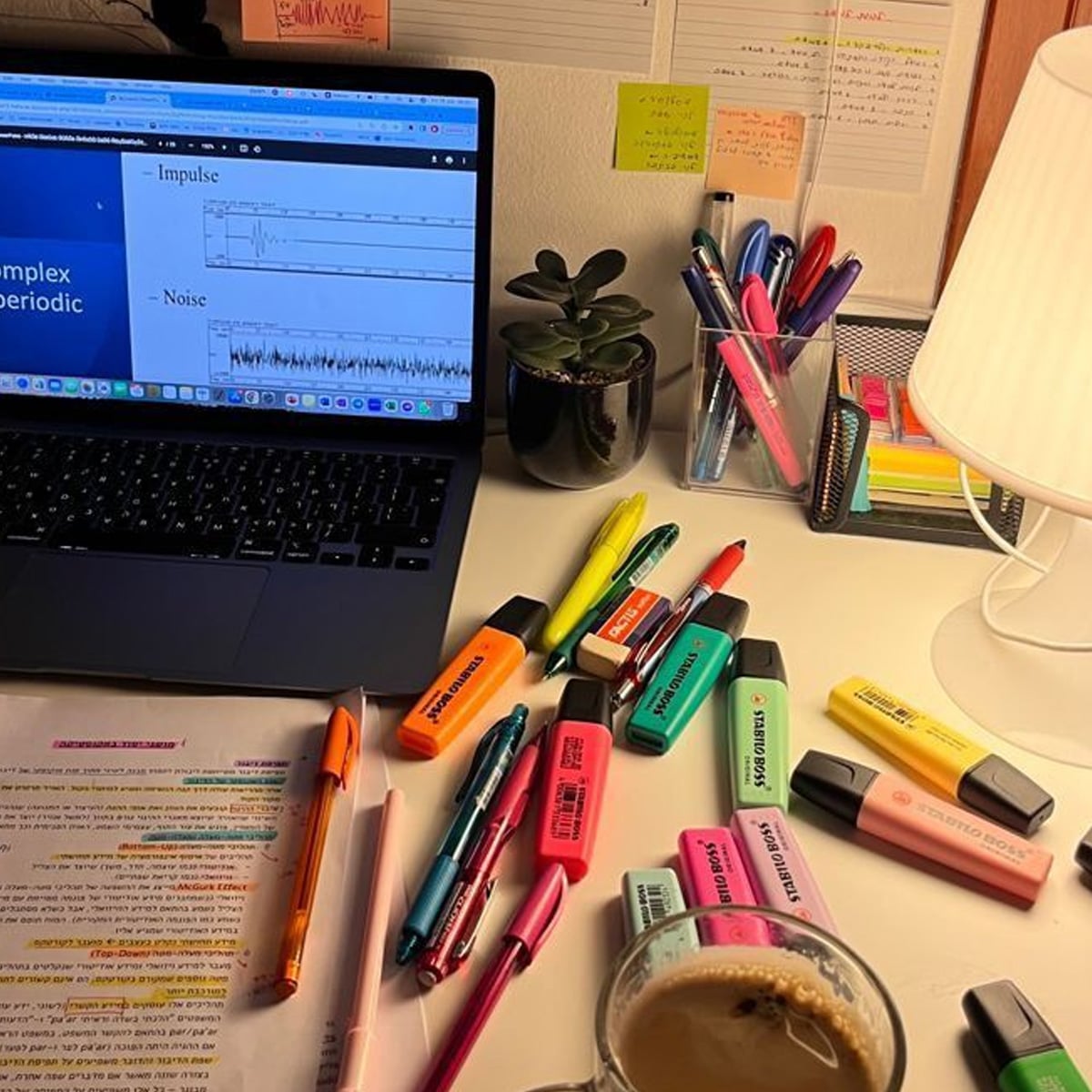 A desk with a laptop showing a graph, many colorful highlighters and pens, a cup of coffee, study notes, a small plant, a lamp, and sticky notes on the wall, creating a busy study or work atmosphere.