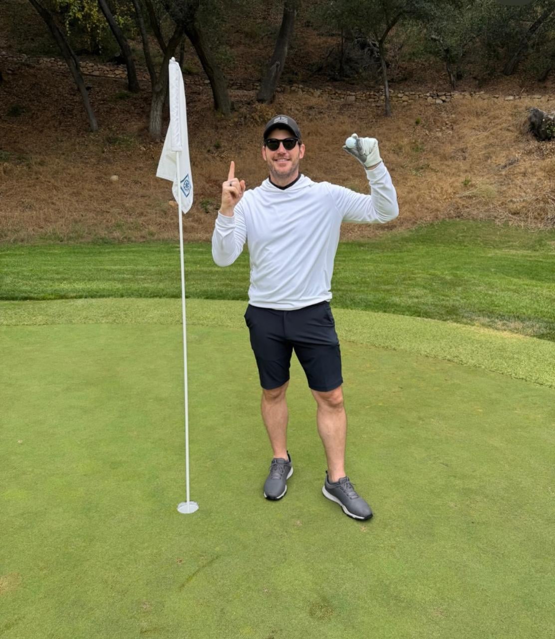 A man wearing a white long-sleeve shirt, black shorts, and a cap stands on a golf green next to a flag, smiling, holding a golf ball in one hand, and raising his index finger with the other. Trees and grass are in the background.