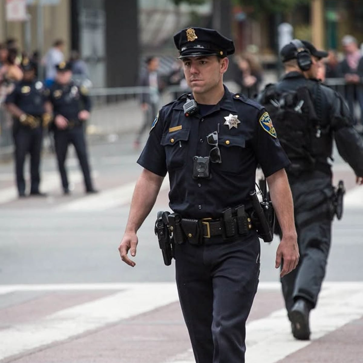 A police officer wearing a uniform and equipment walks across a city street, with other officers and people visible in the background.