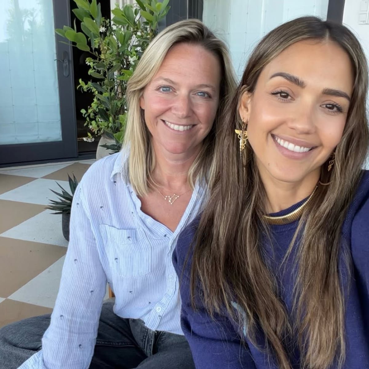 Two women sitting closely together and smiling at the camera. One has long blonde hair and is wearing a light blue shirt; the other has long brown hair and is wearing a dark blue top. There are plants and a checkered floor in the background.