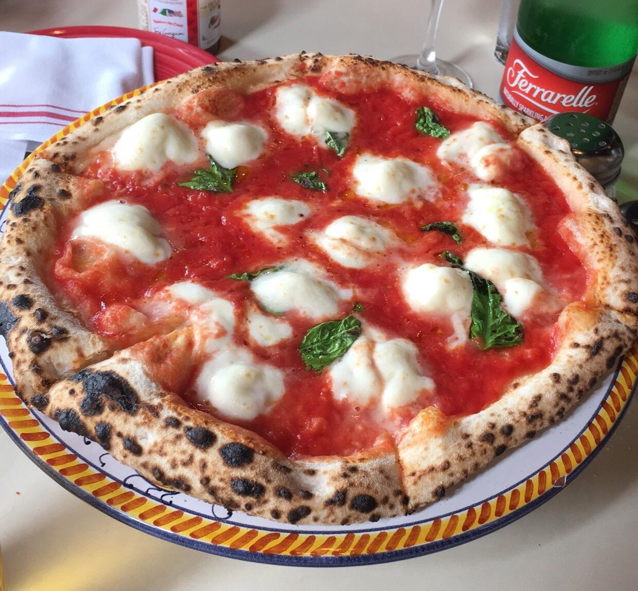 A Neapolitan-style Margherita pizza with a charred crust, topped with tomato sauce, melted mozzarella cheese, and fresh basil leaves, served on a colorful plate. A bottle of sparkling water is visible in the background.