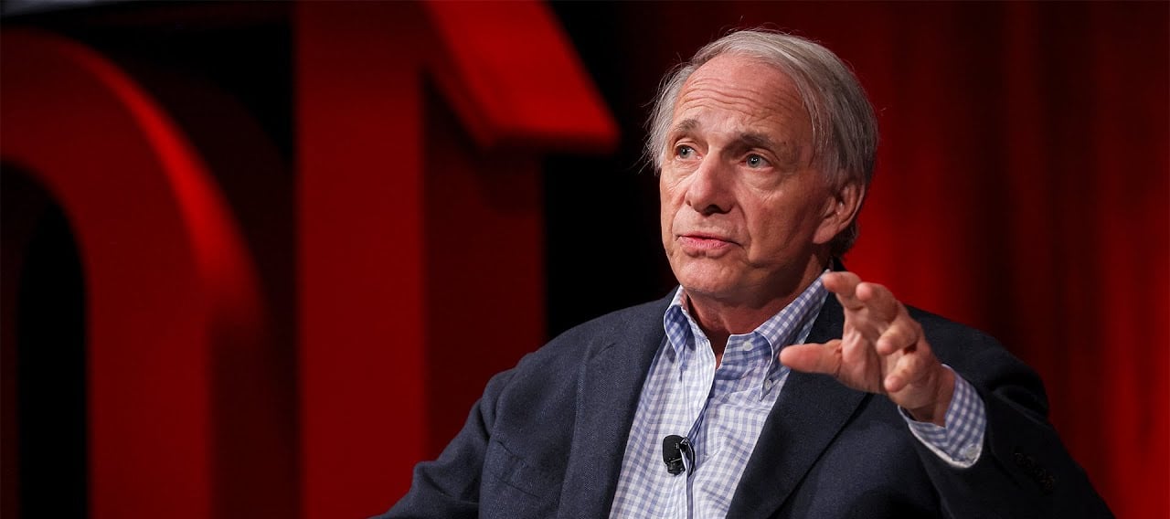 A man with gray hair, wearing a dark blazer and checkered shirt, speaks while gesturing with his hand. He is seated on a stage with red curtains and large red letters in the background.