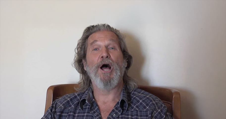 A bearded man with long, gray hair sits on a wooden chair against a plain light-colored wall, wearing a plaid shirt and looking directly at the camera with his mouth open as if speaking or singing.