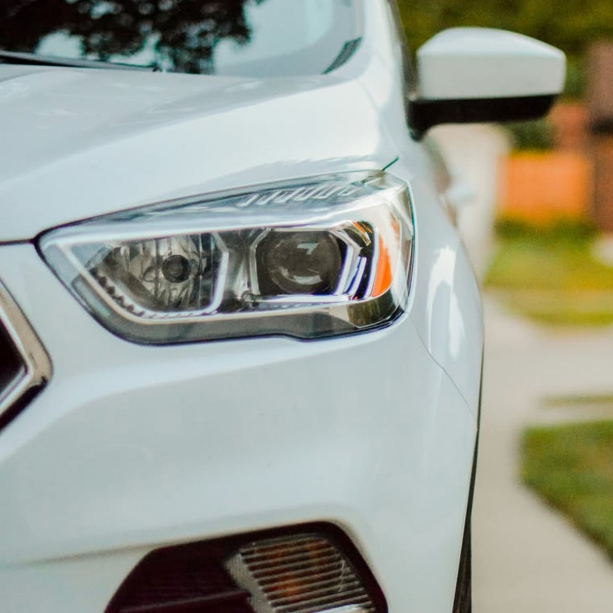 Close-up of the front left headlight and side mirror of a white car parked outdoors, with a blurred background of greenery and a sidewalk.