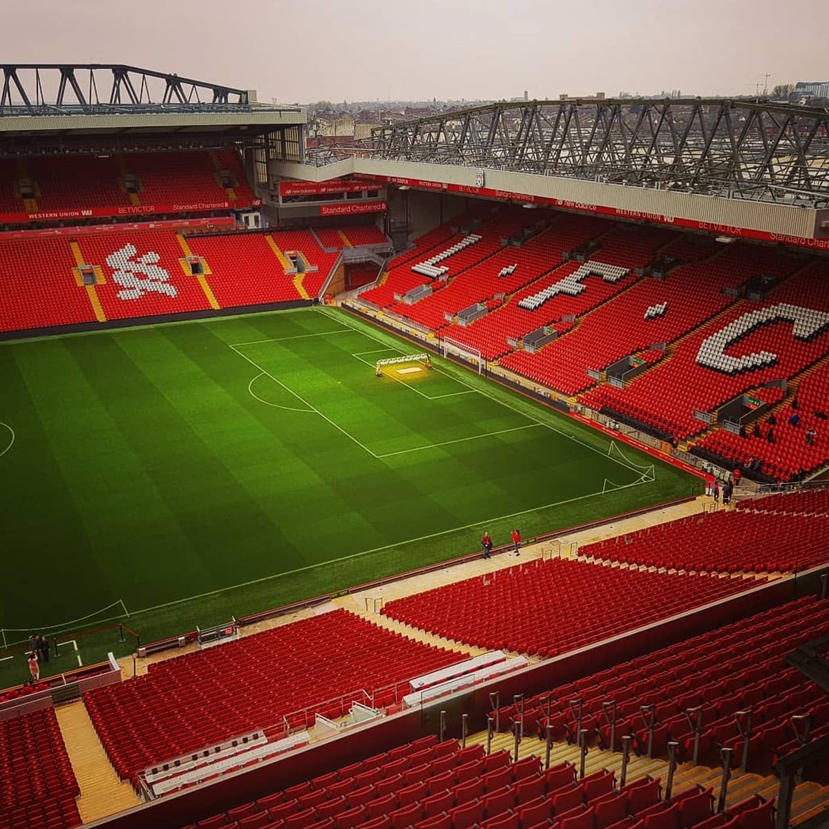 View of a large, empty football stadium with red seats spelling "L F C" and white liver bird logos; a well-kept green pitch is in the center, and a few people are visible near the field.