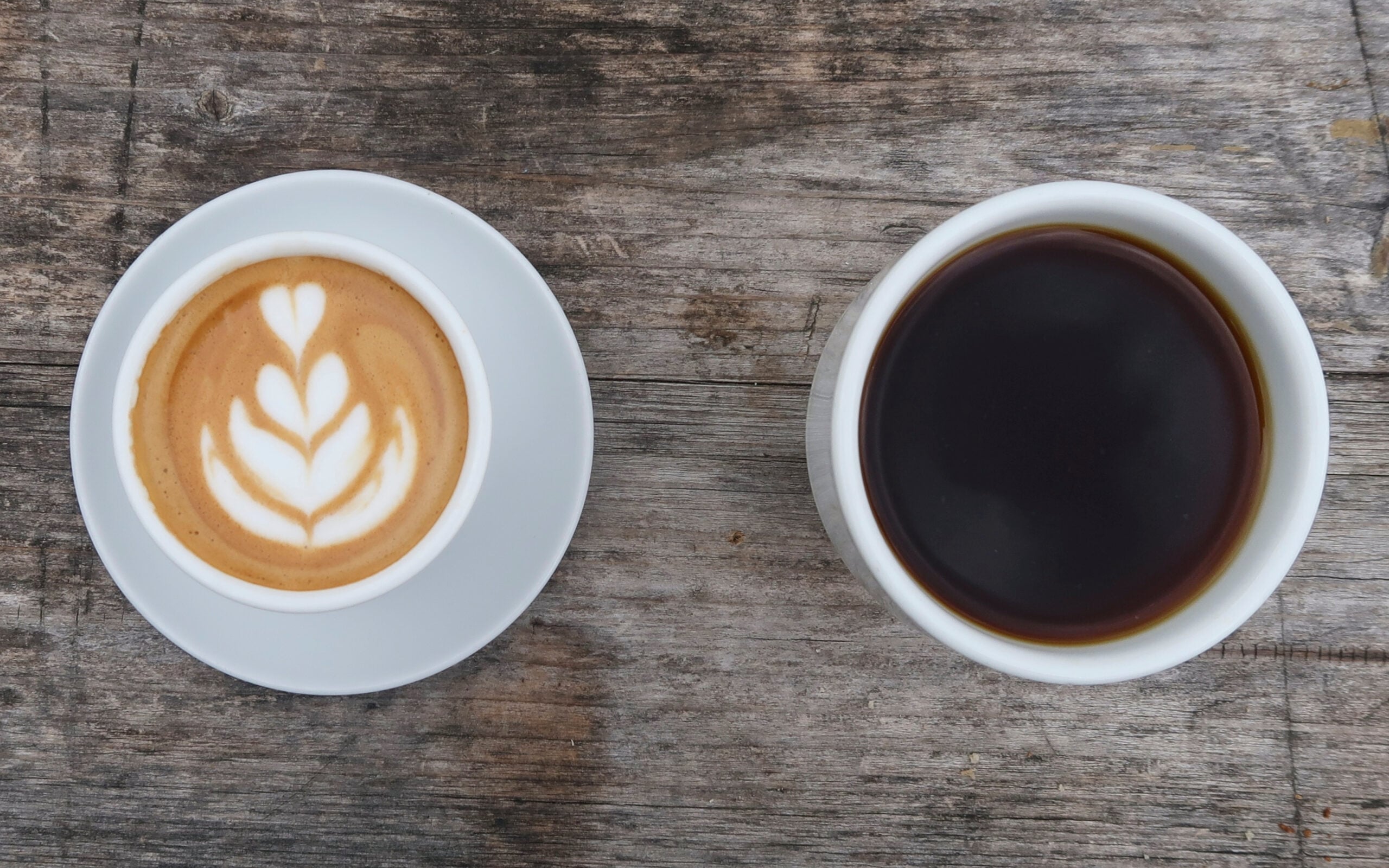 Two white cups on saucers, one with a latte featuring leaf-shaped foam art, the other with black coffee, both placed on a rustic wooden table.