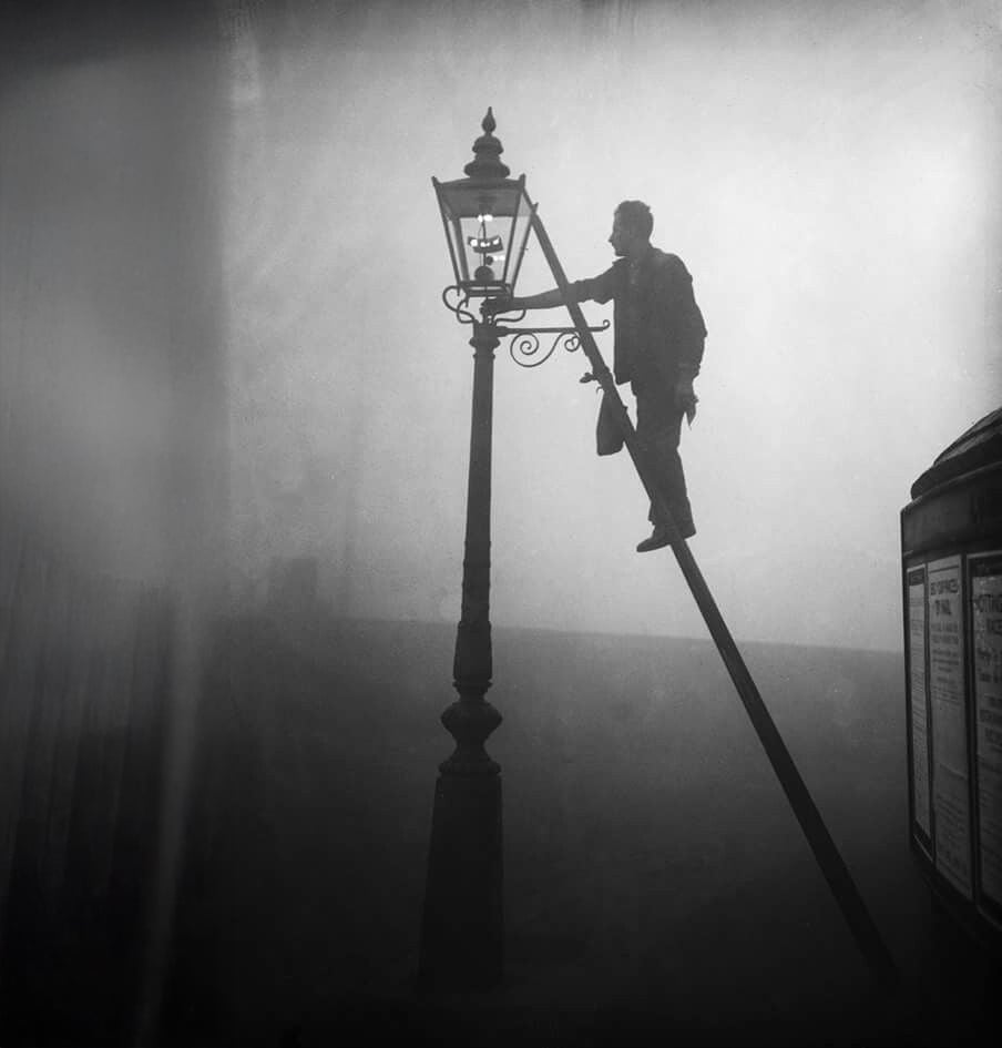A person stands on a ladder, lighting a gas street lamp in thick fog; the figure and lamp are silhouetted against the misty background, creating a moody, atmospheric scene.