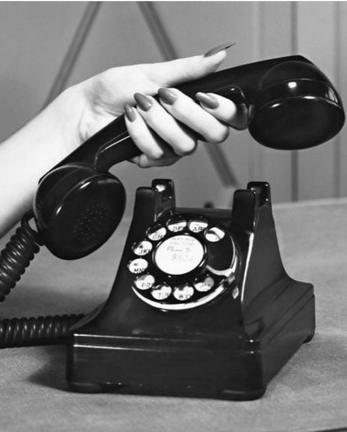 A hand with painted nails lifts the receiver of a vintage rotary dial telephone placed on a table. The image is in black and white.