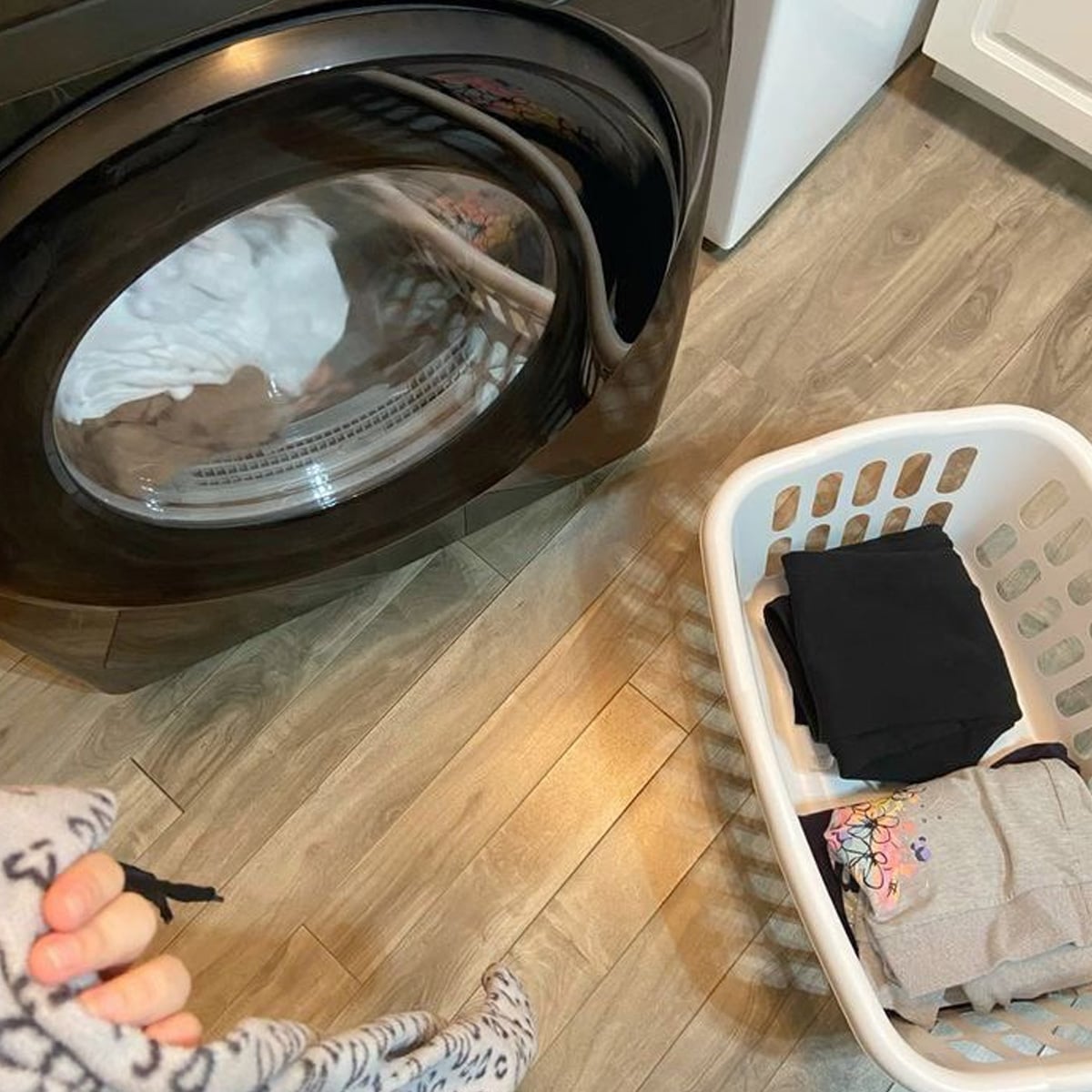 A person holds laundry near a front-loading washing machine. Next to the machine is a white laundry basket containing folded clothes on a wood-patterned floor.