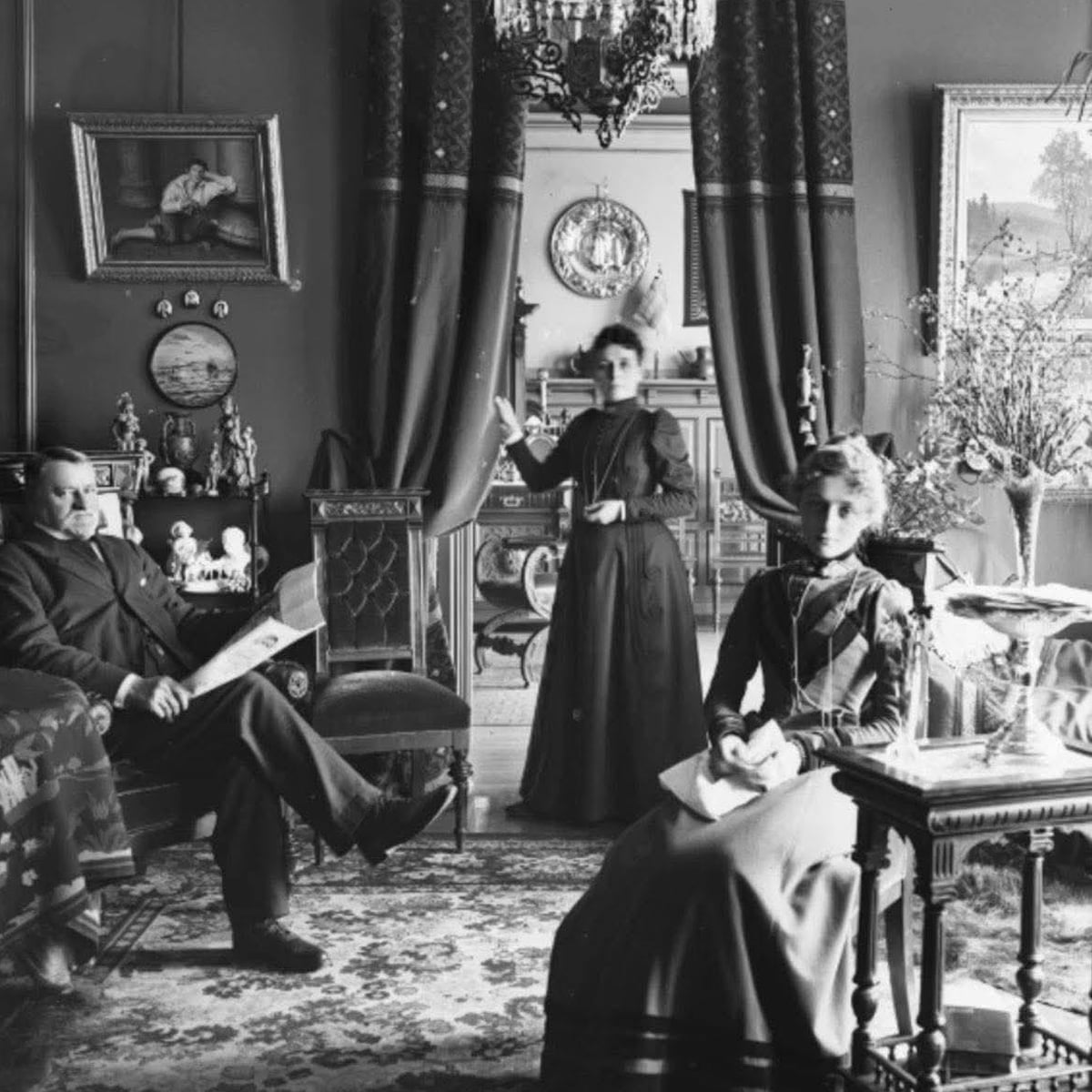 Black-and-white photo of three people in a Victorian-style parlor: a seated man reading a newspaper, and two women, one standing and one sitting, surrounded by ornate furniture, framed art, and decorative items.