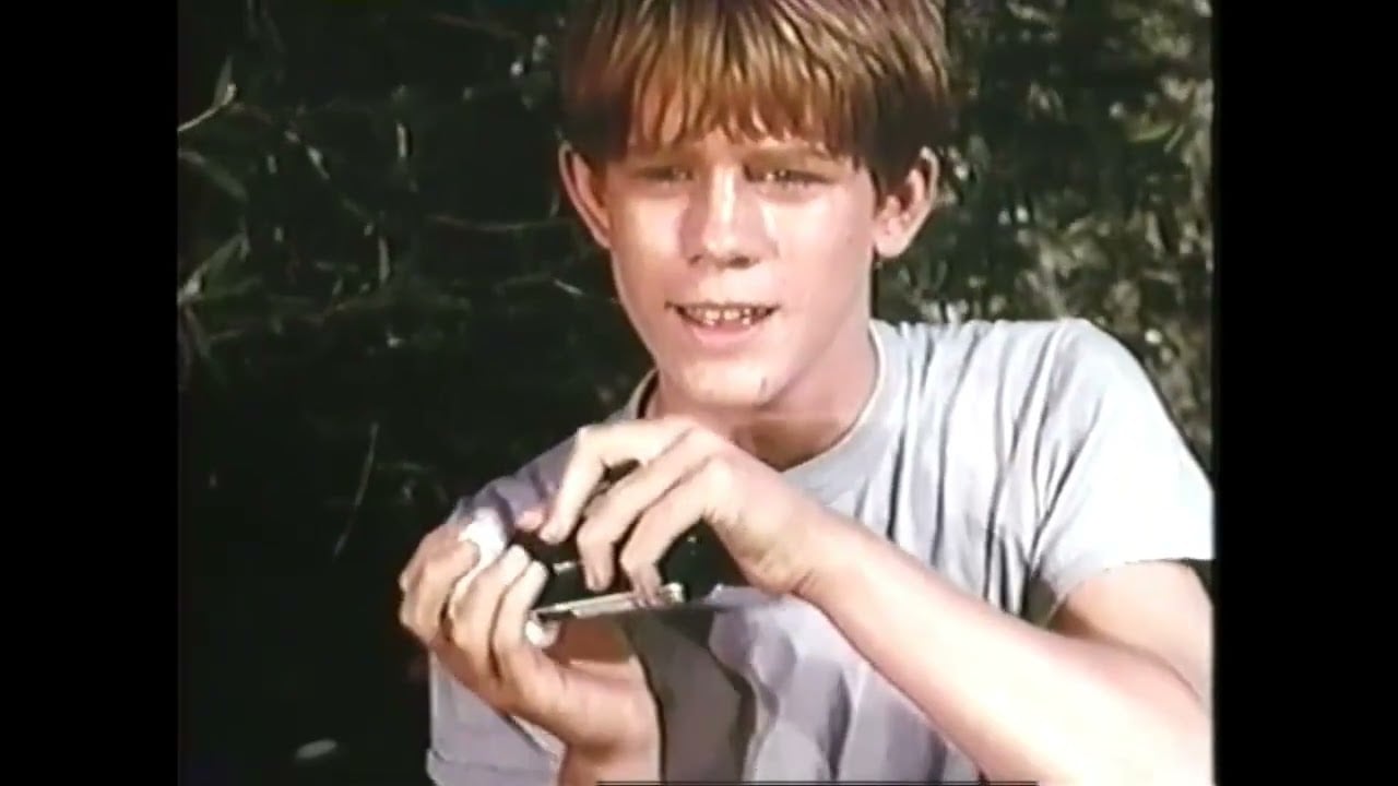 A teenage boy with reddish-brown hair and a light gray T-shirt is smiling while holding and operating a small black camera outdoors, with greenery in the background.