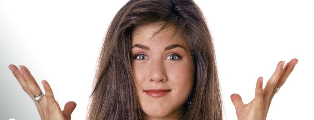 A woman with long brown hair smiles slightly and raises both hands in a shrugging gesture against a plain white background. She is wearing rings and has natural makeup.