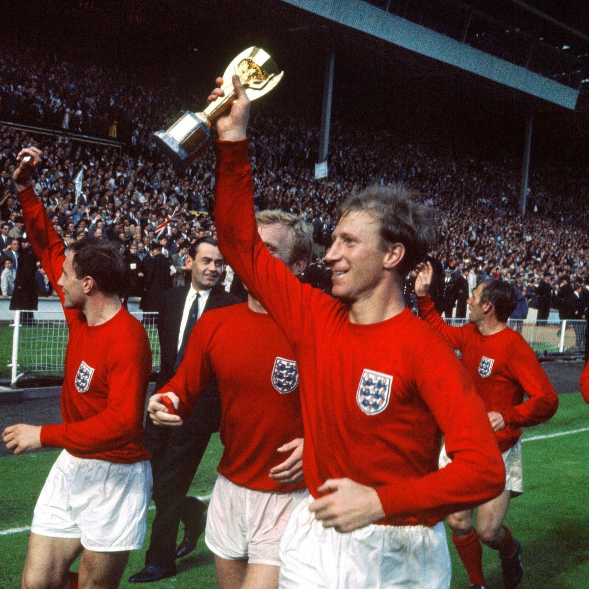 England football players in red jerseys celebrate on the field, one holding the FIFA World Cup trophy aloft, with cheering fans in a crowded stadium in the background.