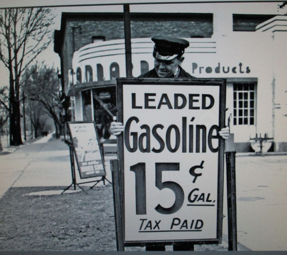 A man in a uniform holds a large sign reading "Leaded Gasoline 15¢ Gal. Tax Paid" on a sidewalk near a vintage gas station. The scene appears to be from an earlier era, with old-fashioned buildings in the background.