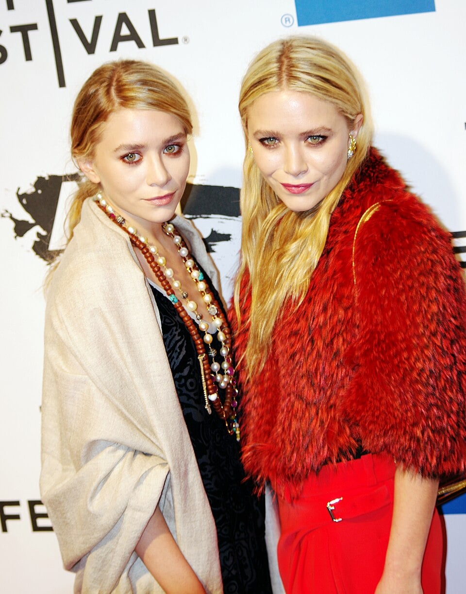 Two women with long blonde hair stand together at an event, both wearing stylish outfits—one in a tan shawl with layered necklaces, the other in a red textured jacket. A step-and-repeat backdrop is behind them.