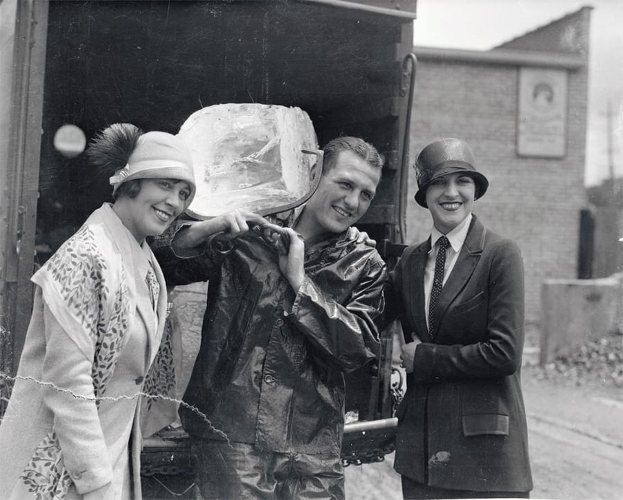 Three people pose in front of an open truck. A man in a raincoat carries a large block of ice on his shoulder, smiling, while two women in 1920s-style clothing and hats stand beside him, also smiling at the camera.