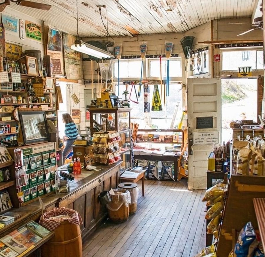 A cozy, old-fashioned general store with wooden floors, shelves stocked with snacks and goods, vintage posters on the walls, and sunlight streaming through large front windows. A person stands behind the counter.