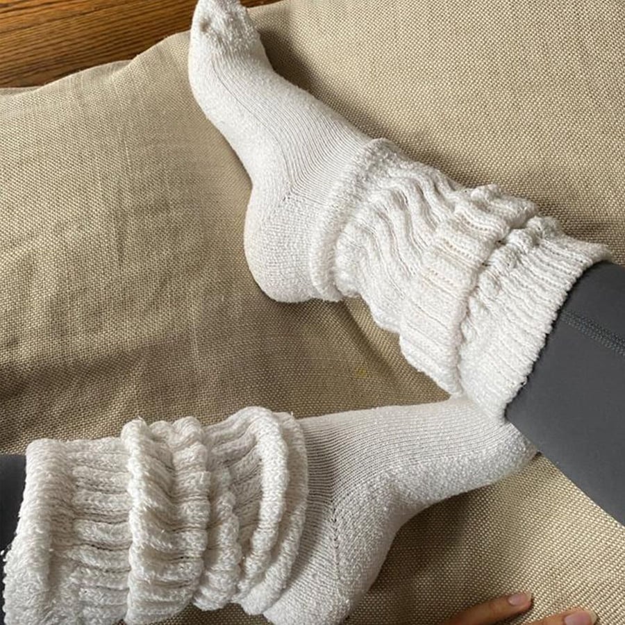 Two feet wearing thick, white, ruffled socks rest on a beige cushion. The person is wearing dark gray leggings, and the wooden floor is partially visible in the background.