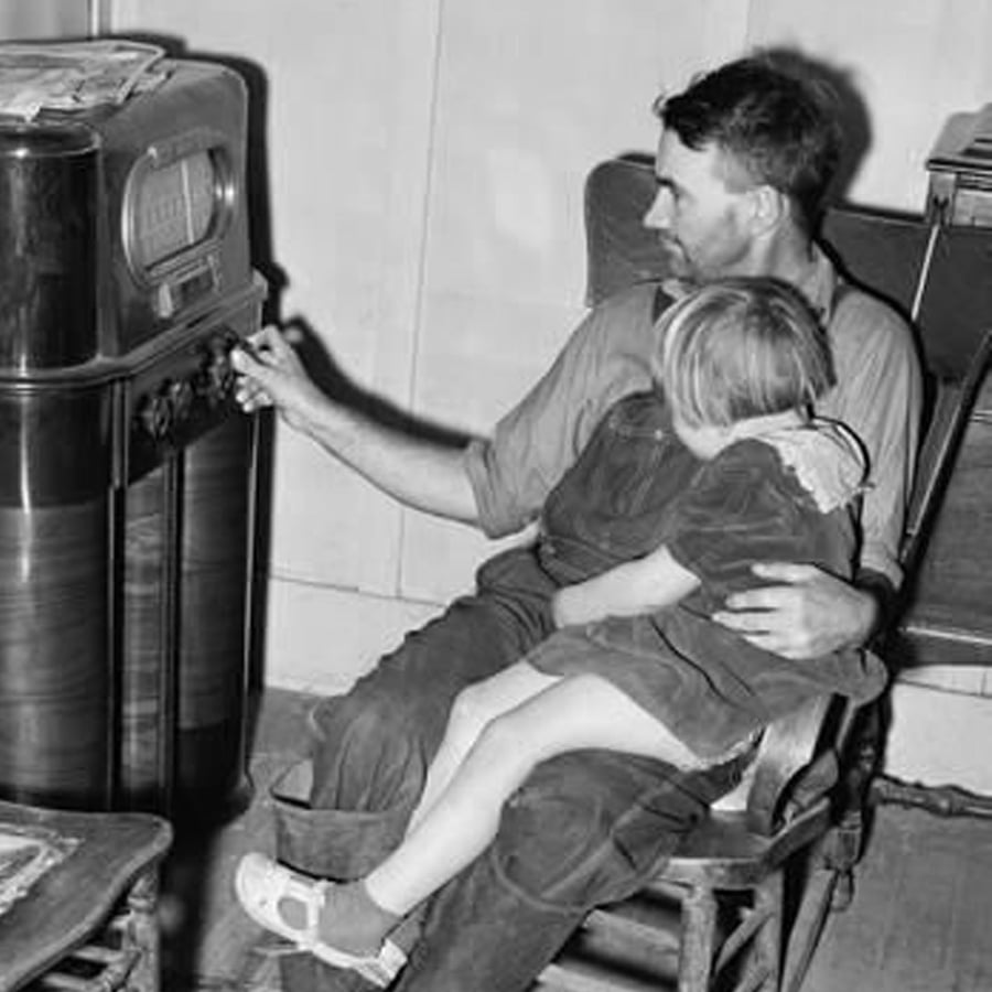 A man sits in a wooden chair, holding a young girl on his lap, as he tunes a large vintage radio. The child rests her head on his shoulder. Newspapers are stacked on a nearby table.