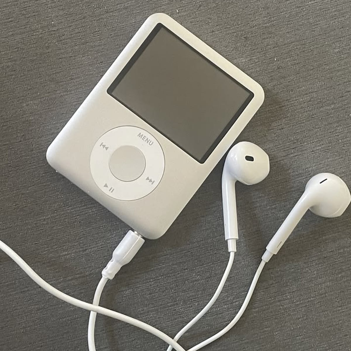A white MP3 player with a circular control wheel and a blank screen is connected to a pair of white wired earbuds, resting on a gray surface.