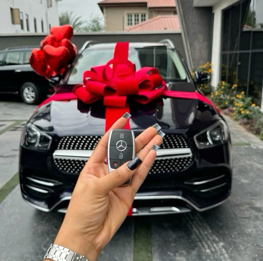 A hand with manicured nails holds a Mercedes-Benz car key fob in front of a black Mercedes-Benz SUV decorated with a large red bow and balloons, suggesting it is a gift.