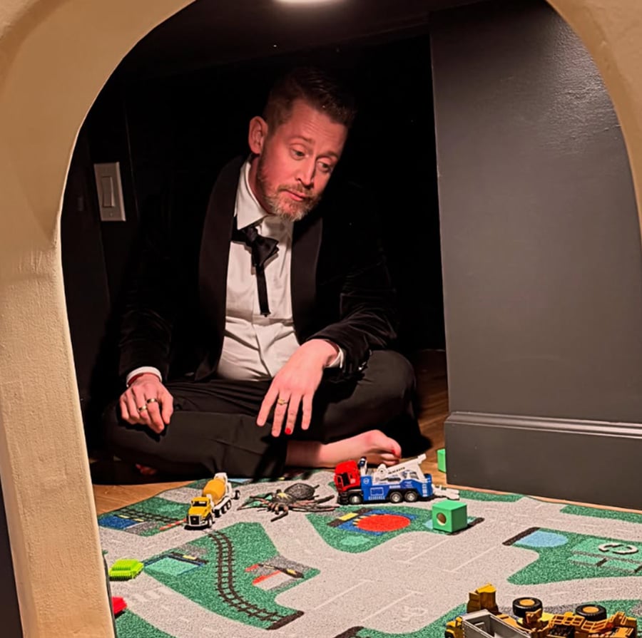 A man in a tuxedo sits cross-legged in a small, dimly lit room, playing with toy trucks and cars on a colorful play mat. He appears thoughtful as he looks at the toys.