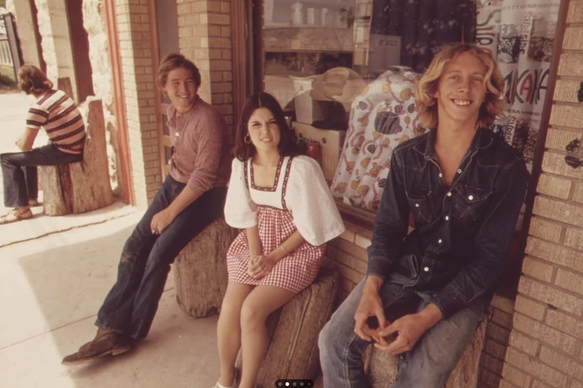 Three young people sit on wooden stumps outside a brick storefront. Two men in casual 1970s clothing and a woman in a red gingham dress smile at the camera. Another person sits in the background, turned away.