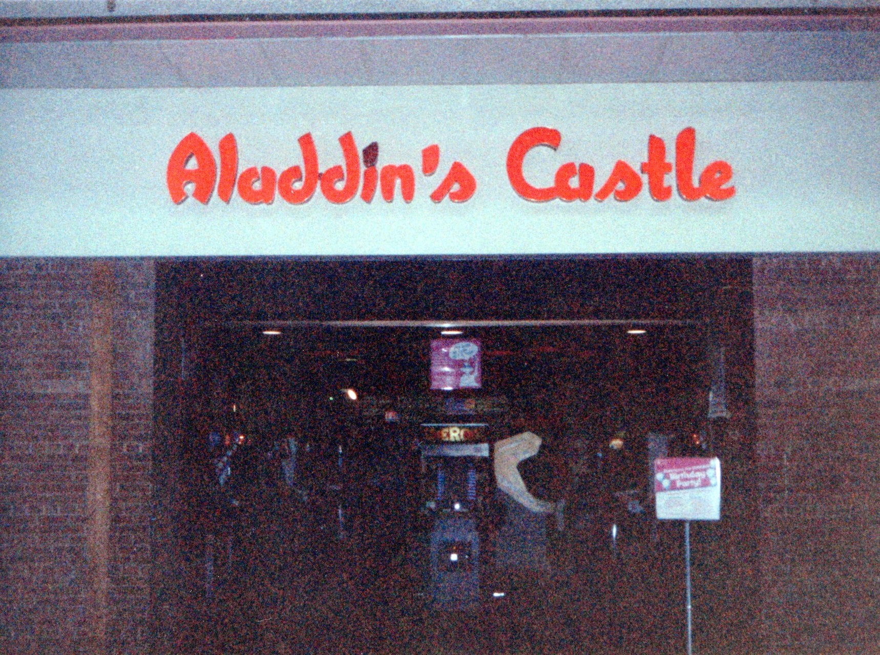 The entrance to an arcade named "Aladdin's Castle," with red, stylized lettering above a dark interior where arcade machines are visible. A sign with information stands near the doorway.