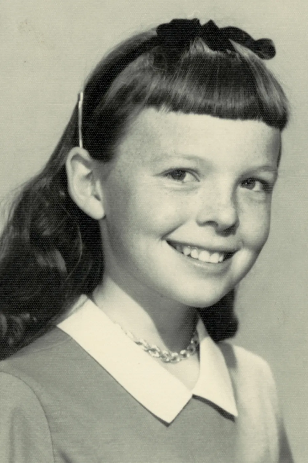 Black-and-white portrait of a young girl smiling, with wavy dark hair, bangs, a headband with a bow, and a collared dress. She wears a beaded necklace and is shown from the shoulders up.