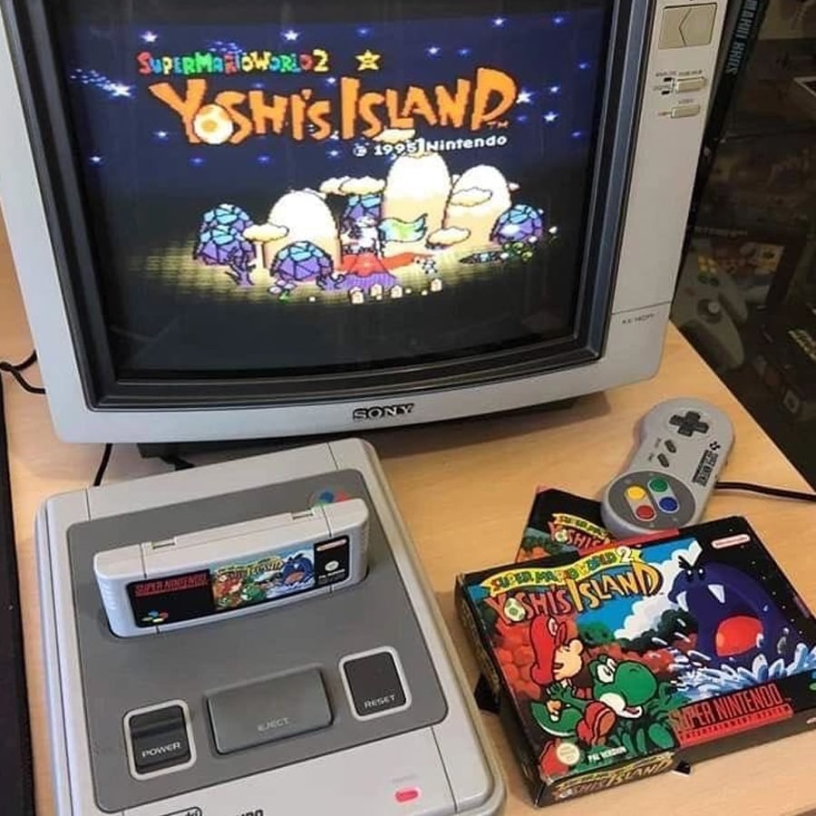A Super Nintendo console with the game "Super Mario World 2: Yoshi's Island" is connected to a CRT TV showing the game's title screen. A controller and the game's box are also visible on the desk.
