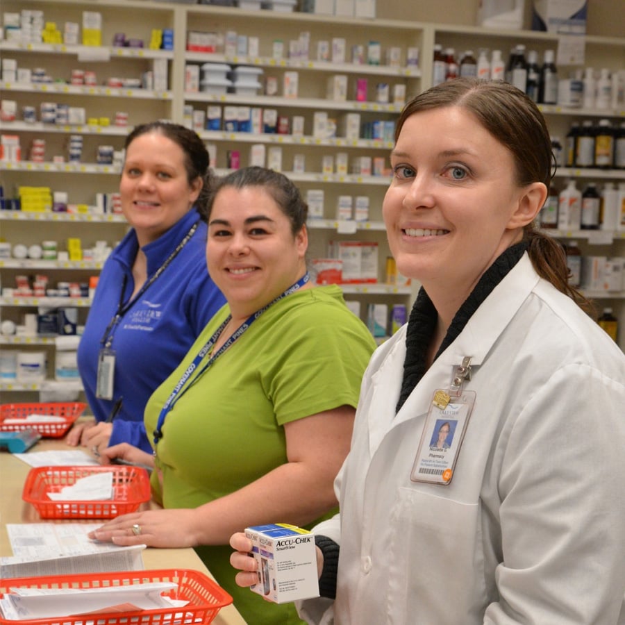Three women in a pharmacy stand behind a counter with red baskets and medication bottles. One wears a white lab coat and holds a box of medicine, while the others wear blue uniforms and smile at the camera. Shelves of medicines are visible.