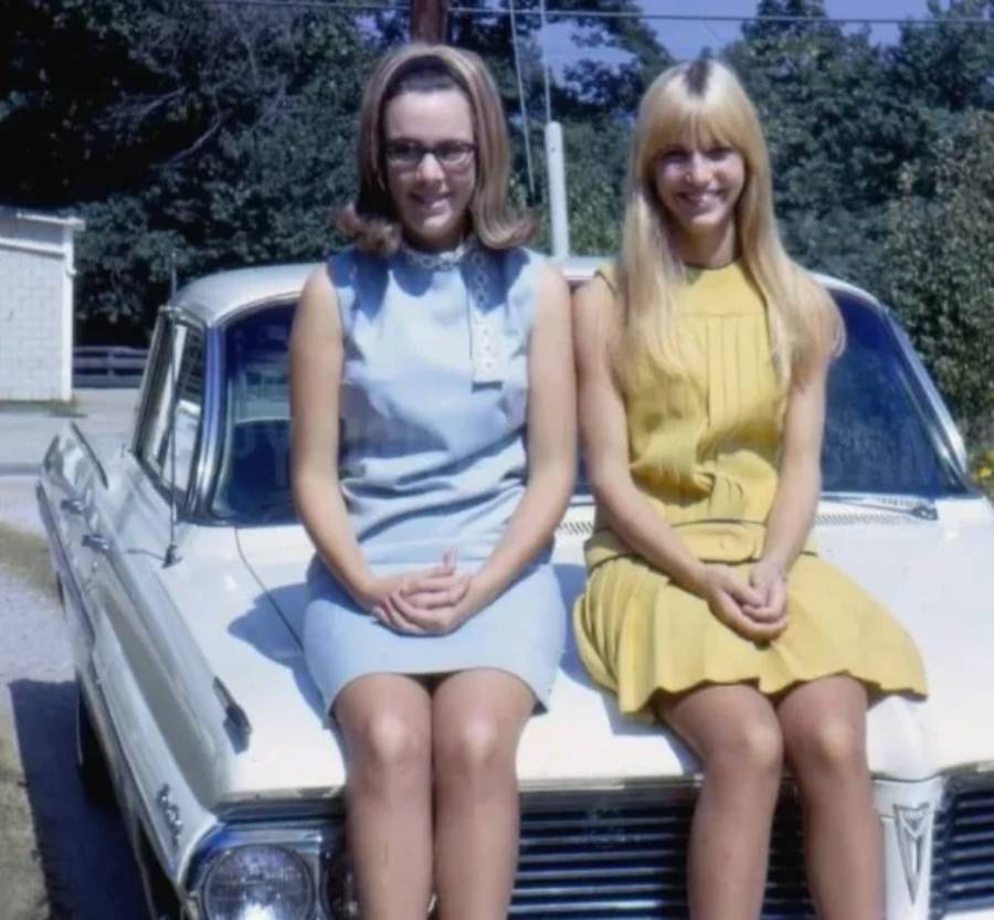Two young women in sleeveless dresses, one in blue and one in yellow, sit side by side on the hood of a vintage white car, smiling at the camera on a sunny day. Trees and a building are visible in the background.