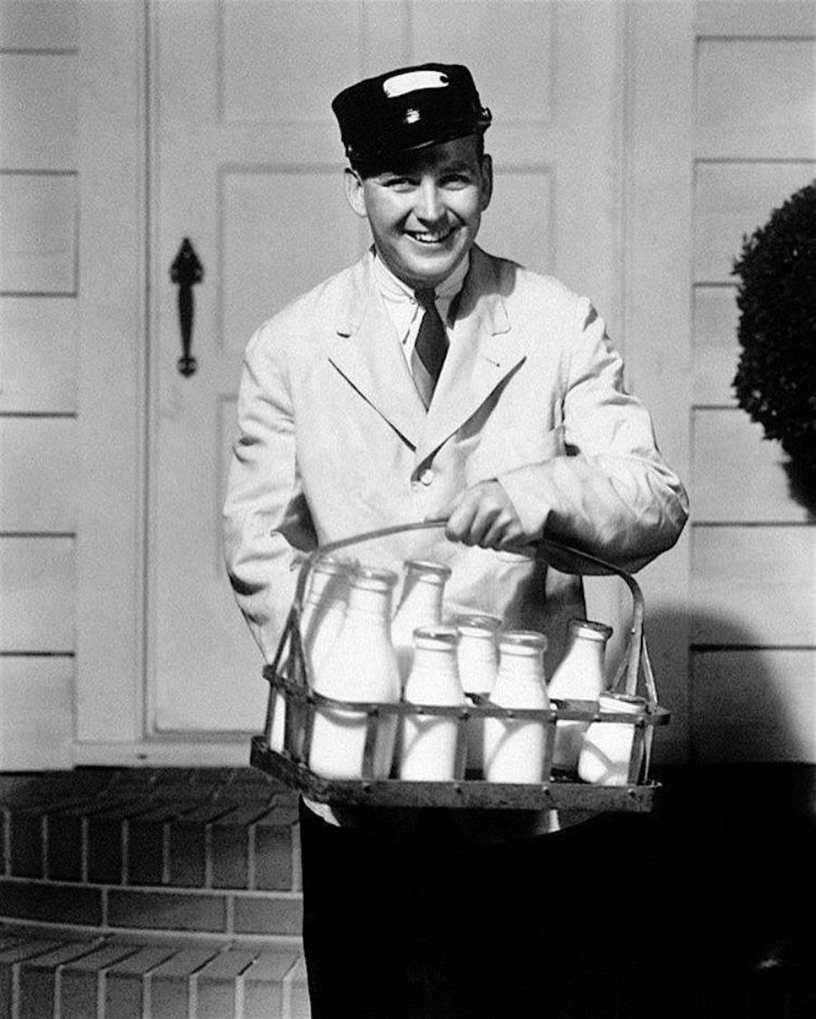 A smiling milkman wearing a cap and uniform holds a basket of glass milk bottles while standing on a doorstep in front of a house.