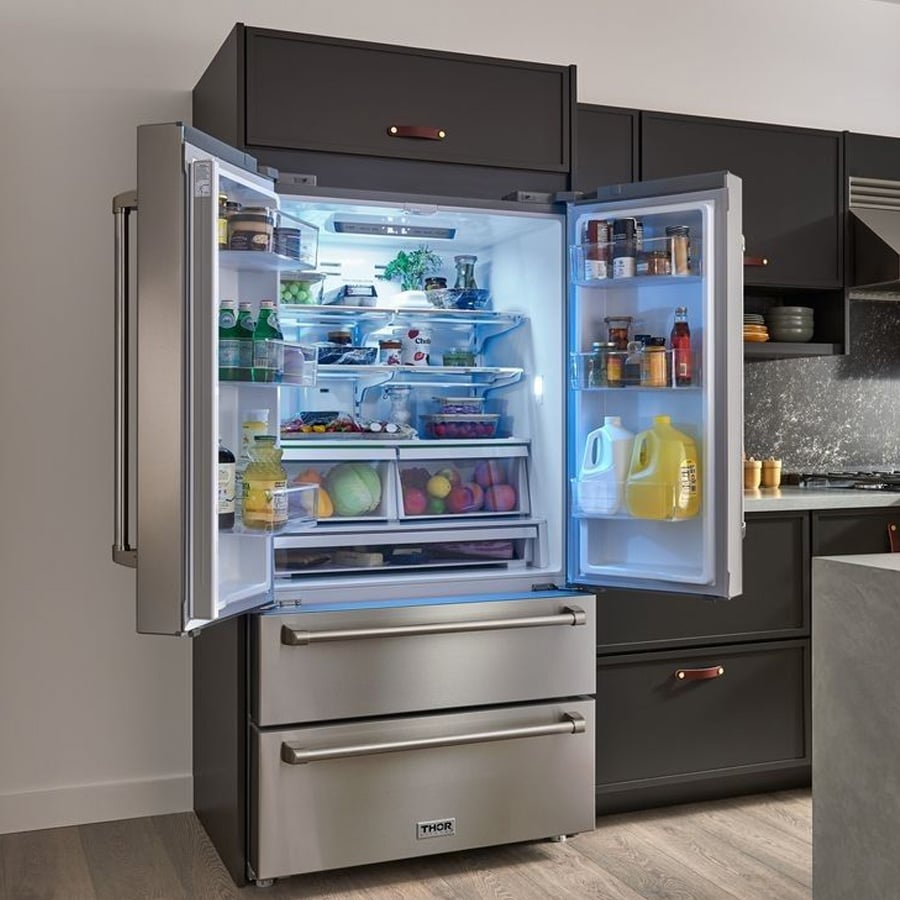 A modern stainless steel refrigerator with French doors and two freezer drawers is open, revealing neatly organized food, drinks, fruits, vegetables, and condiments inside. The kitchen has dark cabinets and wood flooring.
