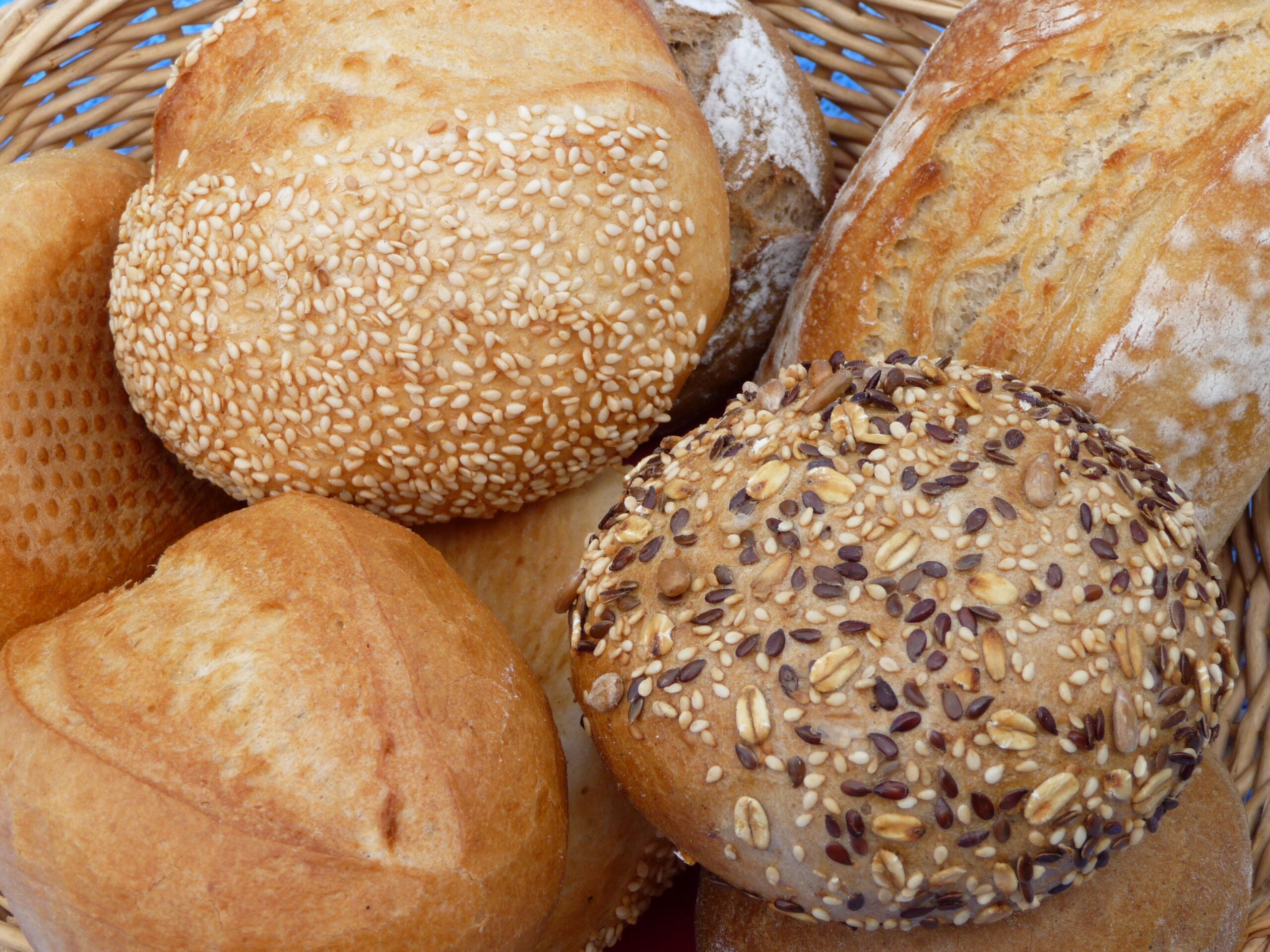 A close-up of assorted bread rolls in a wicker basket, including rolls topped with sesame seeds and mixed seeds, and plain crusty rolls.