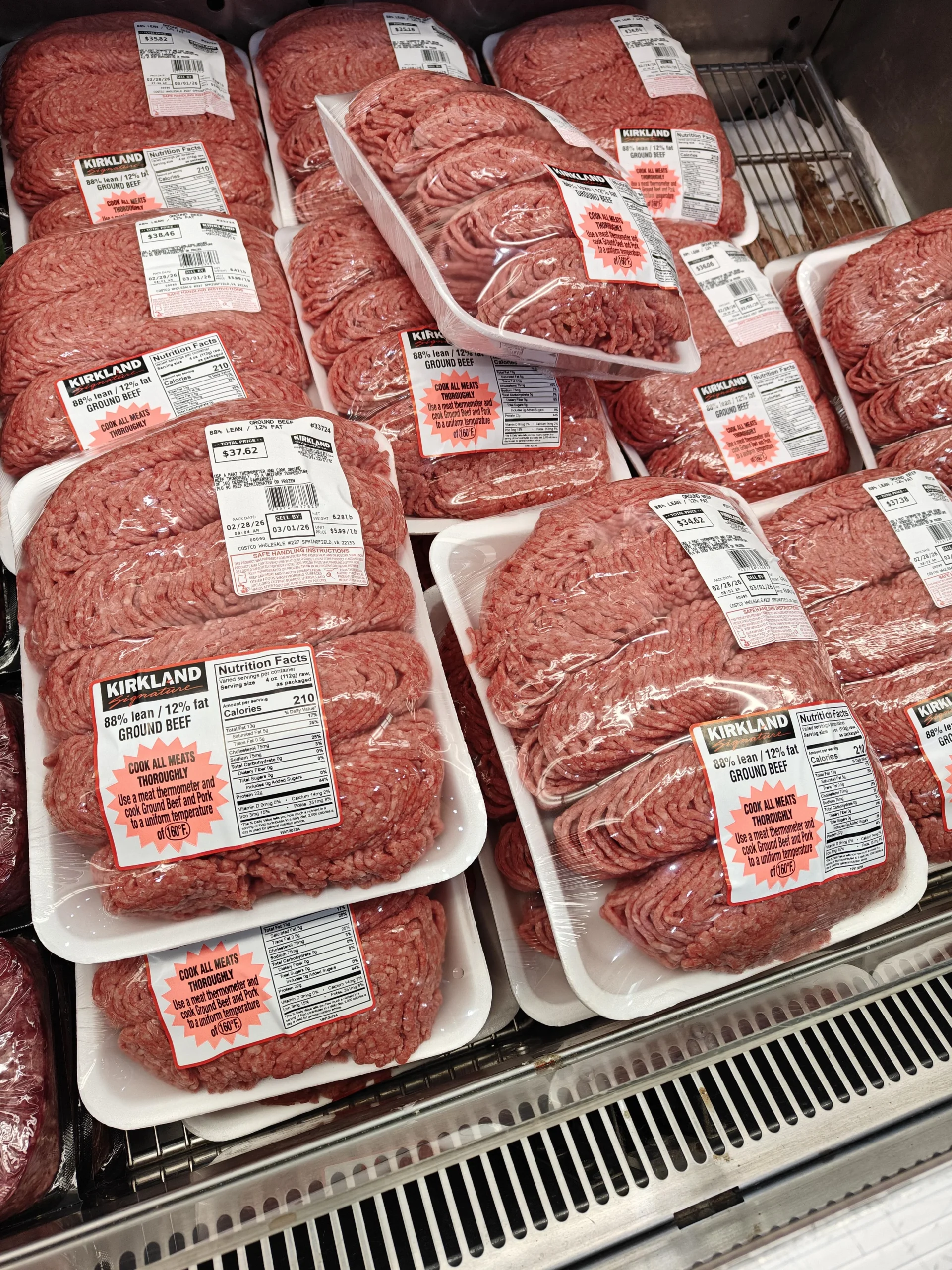 Packages of Kirkland brand ground beef are stacked in a refrigerated display at a store. The meat is packed in clear plastic trays with labels detailing weight, price, and nutritional information.