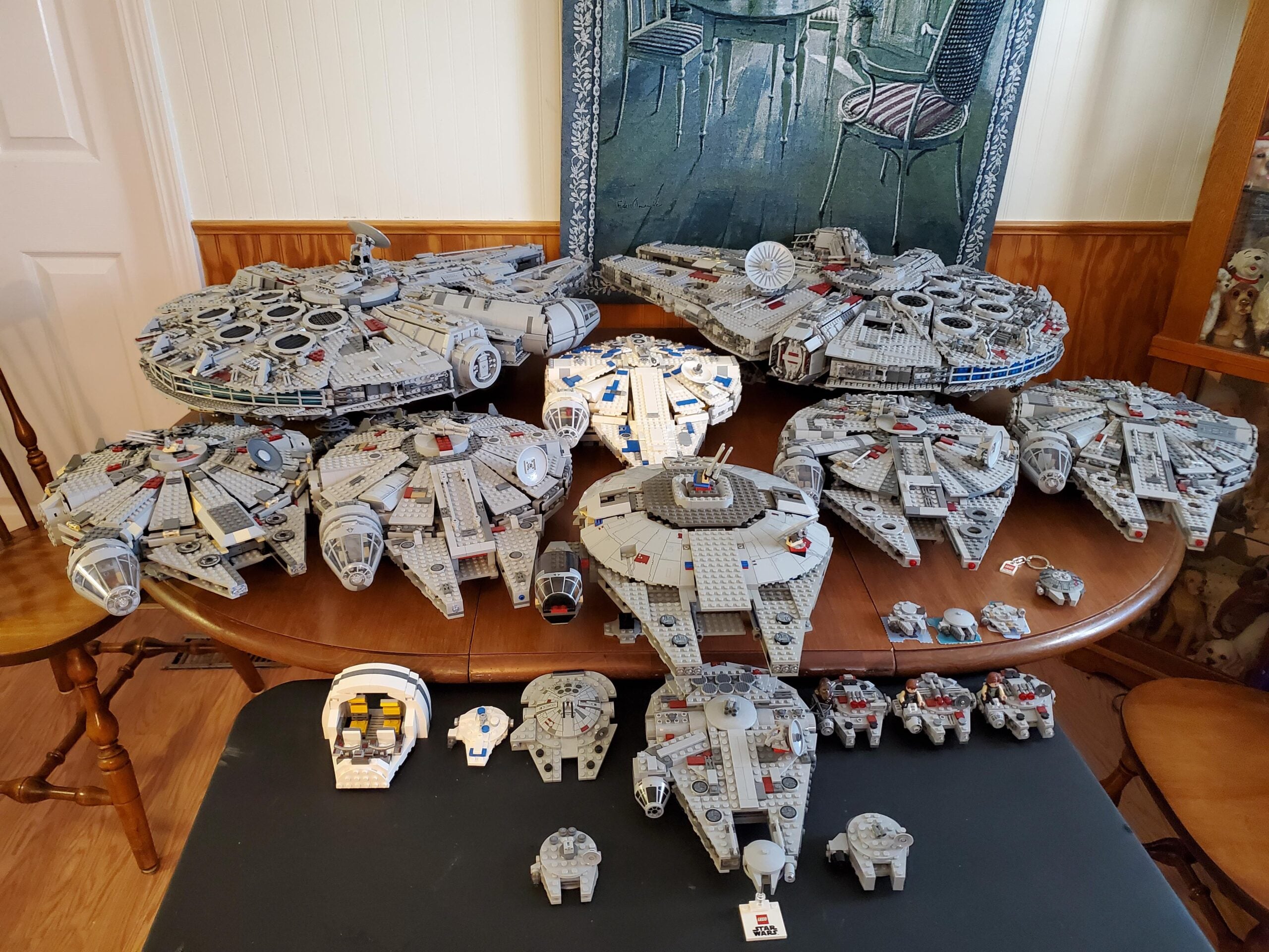 A collection of various LEGO Millennium Falcon models in different sizes are displayed on a table, arranged from largest to smallest, with chairs and home decor in the background.