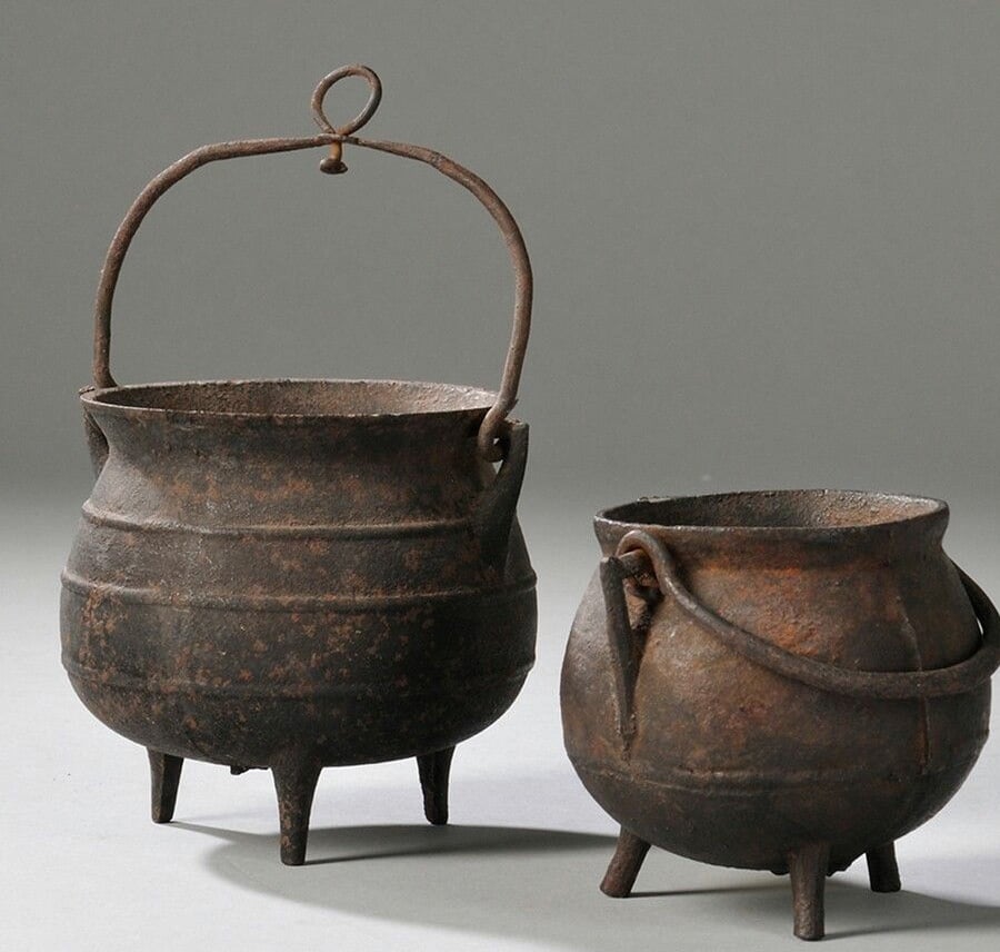 Two old, rusted cast iron cauldrons with three legs each. One is larger with a tall handle and loop, while the other is smaller with a bent handle. Both have a weathered, vintage appearance against a plain background.