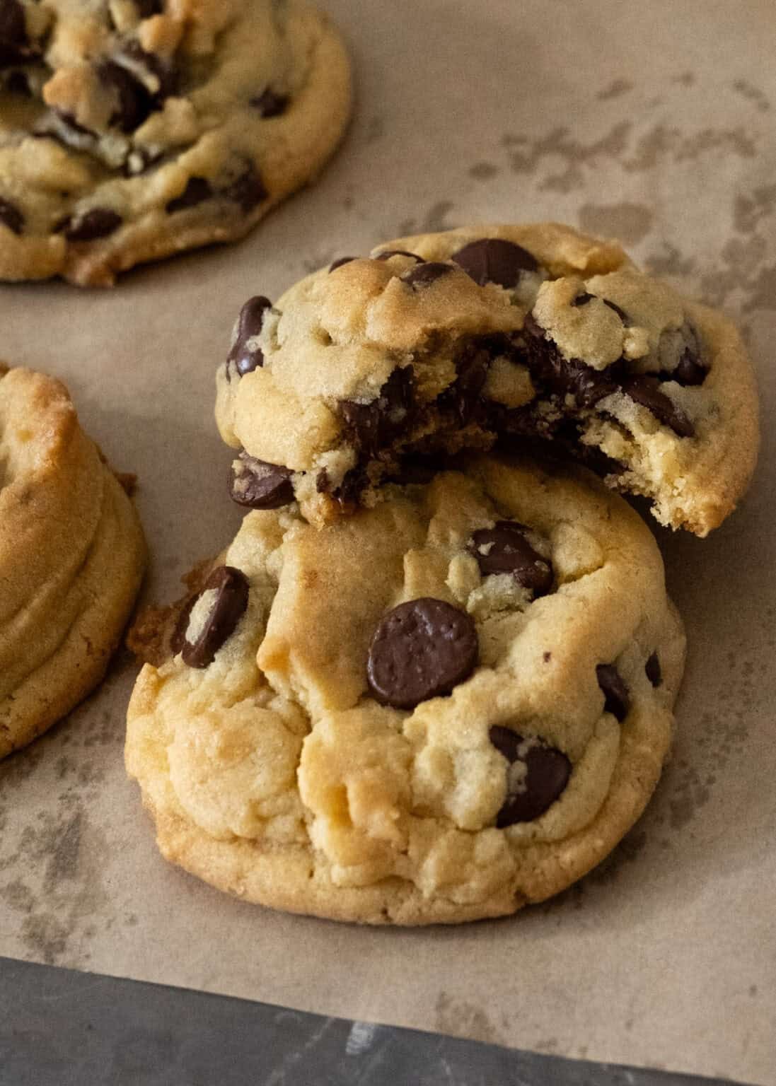 Two thick, soft chocolate chip cookies on parchment paper, with one cookie broken in half to reveal a gooey, chocolate-filled center. Another whole cookie is partially visible in the background.